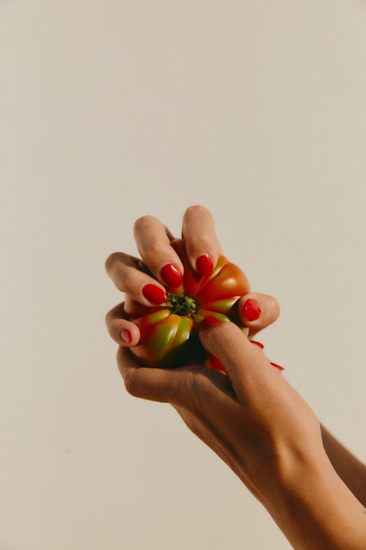 A close-up of a person's hands with bright red nail polish holding a small green and orange tomato against a plain beige background.