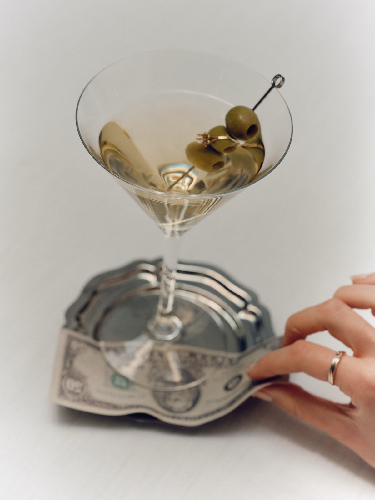 A martini glass filled with a pale golden drink and topped with green olives on a toothpick, resting on a silver tray that is being held by a hand wearing a ring.