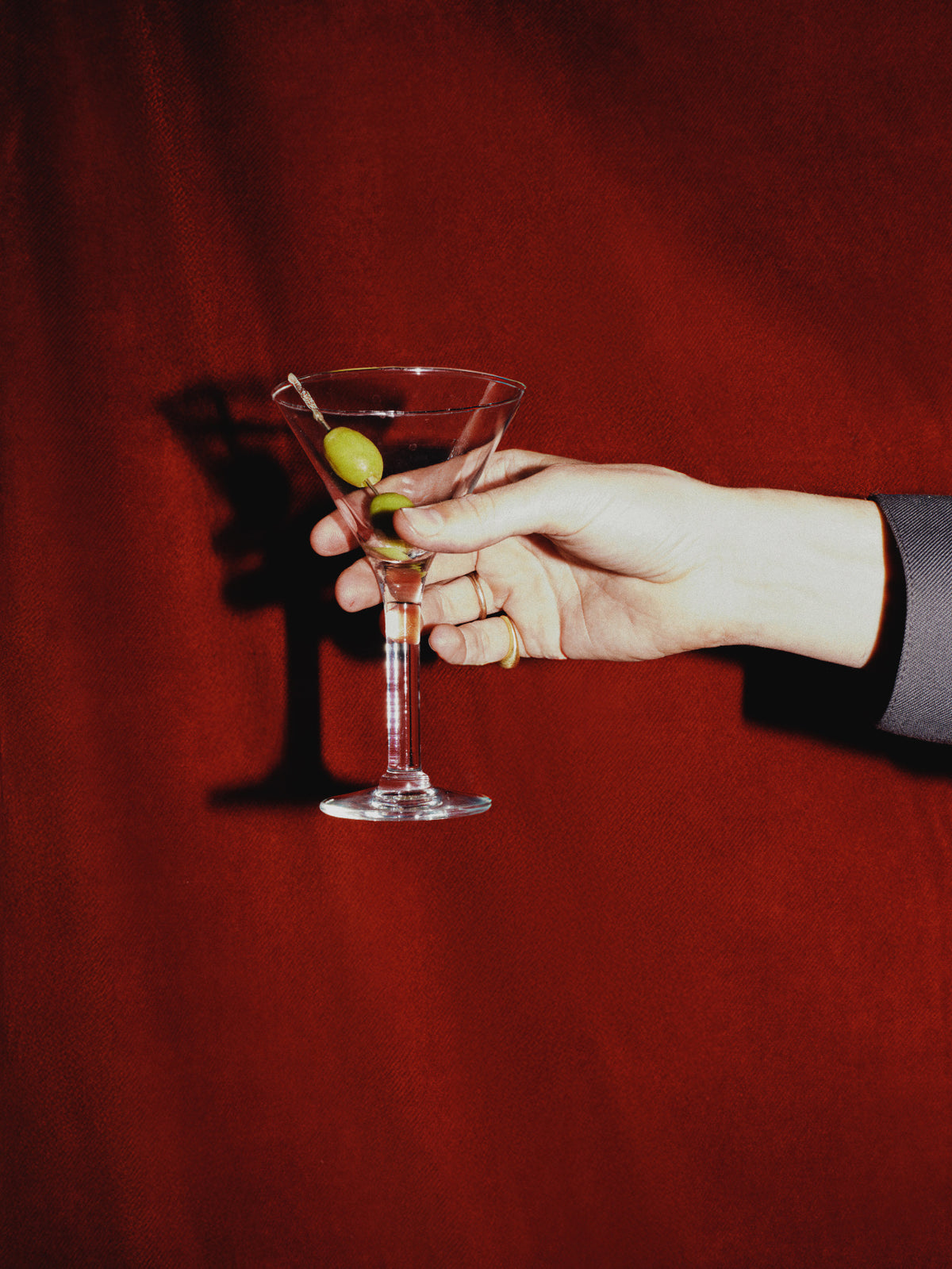 A hand holding a martini glass with olives against a dark red fabric background.