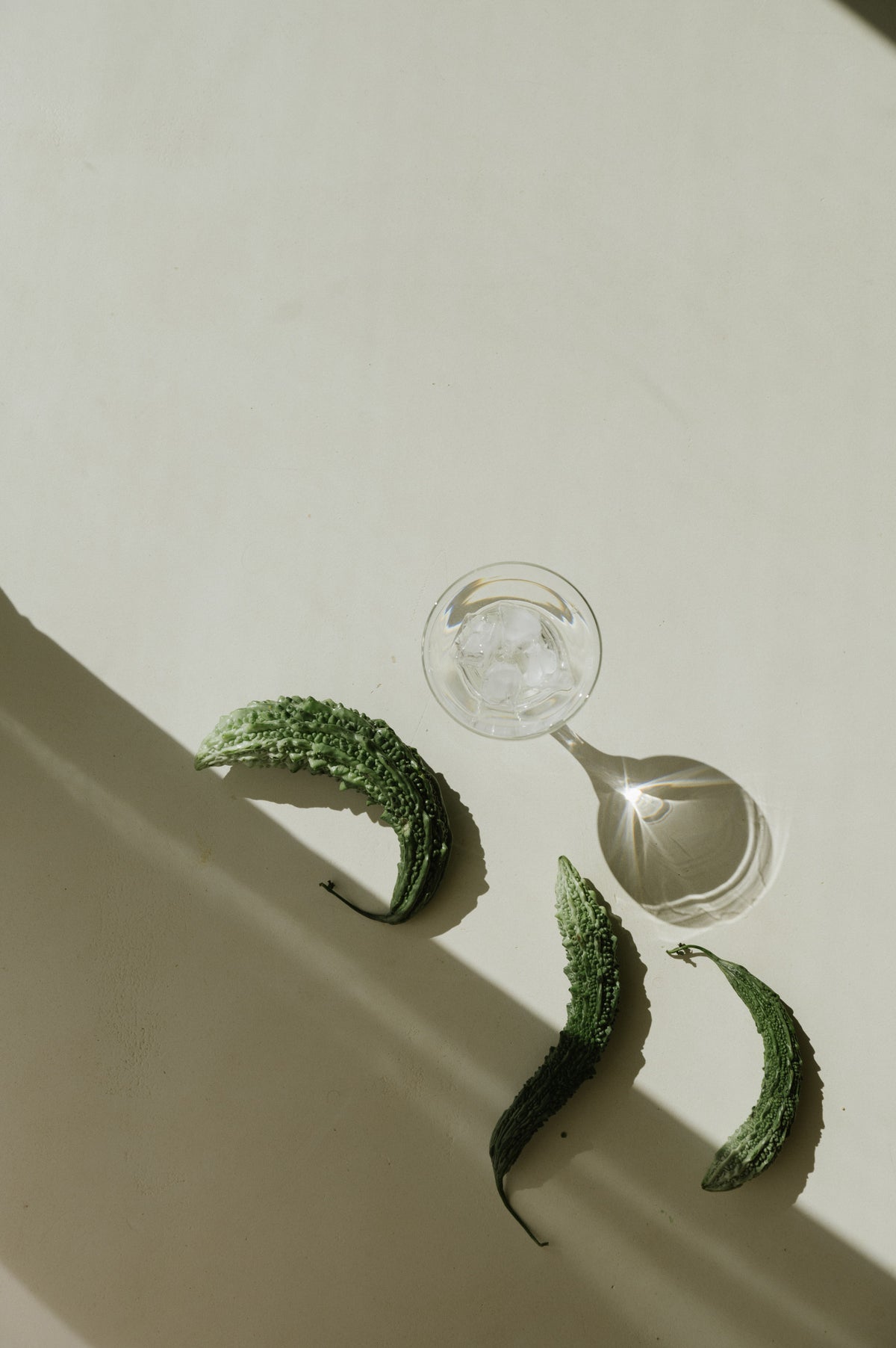 Three green bittersweet melons arranged on a warm cream surface next to a clear glass bowl with ice, cast in sharp diagonal sunlight with strong shadows.