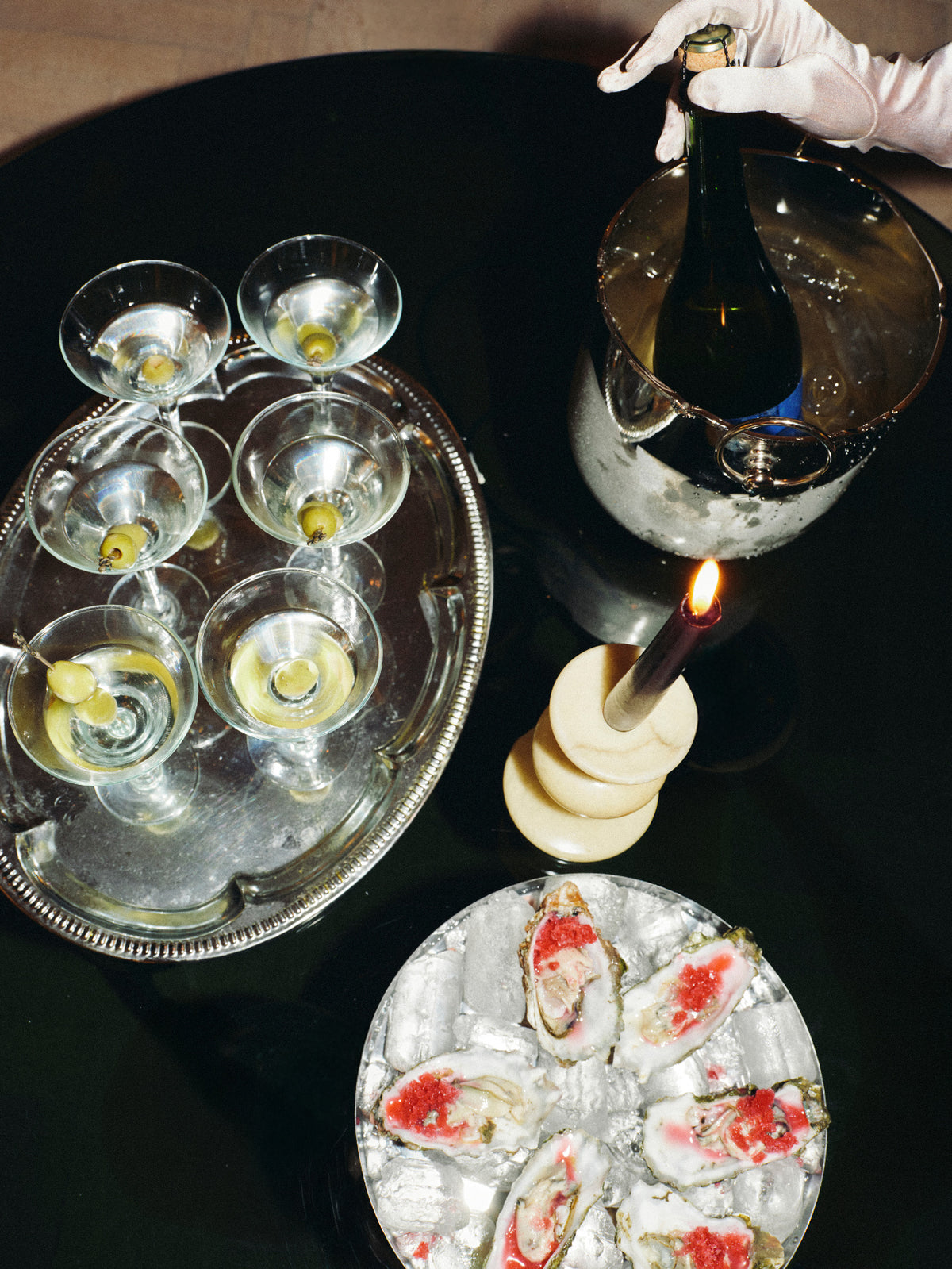 A high-angle view of a luxury bar or catering setup on a black table, featuring a silver tray with six martini glasses garnished with olives, a champagne bucket with a bottle and a gloved hand, a lit candle, and a platter of oysters on ice.