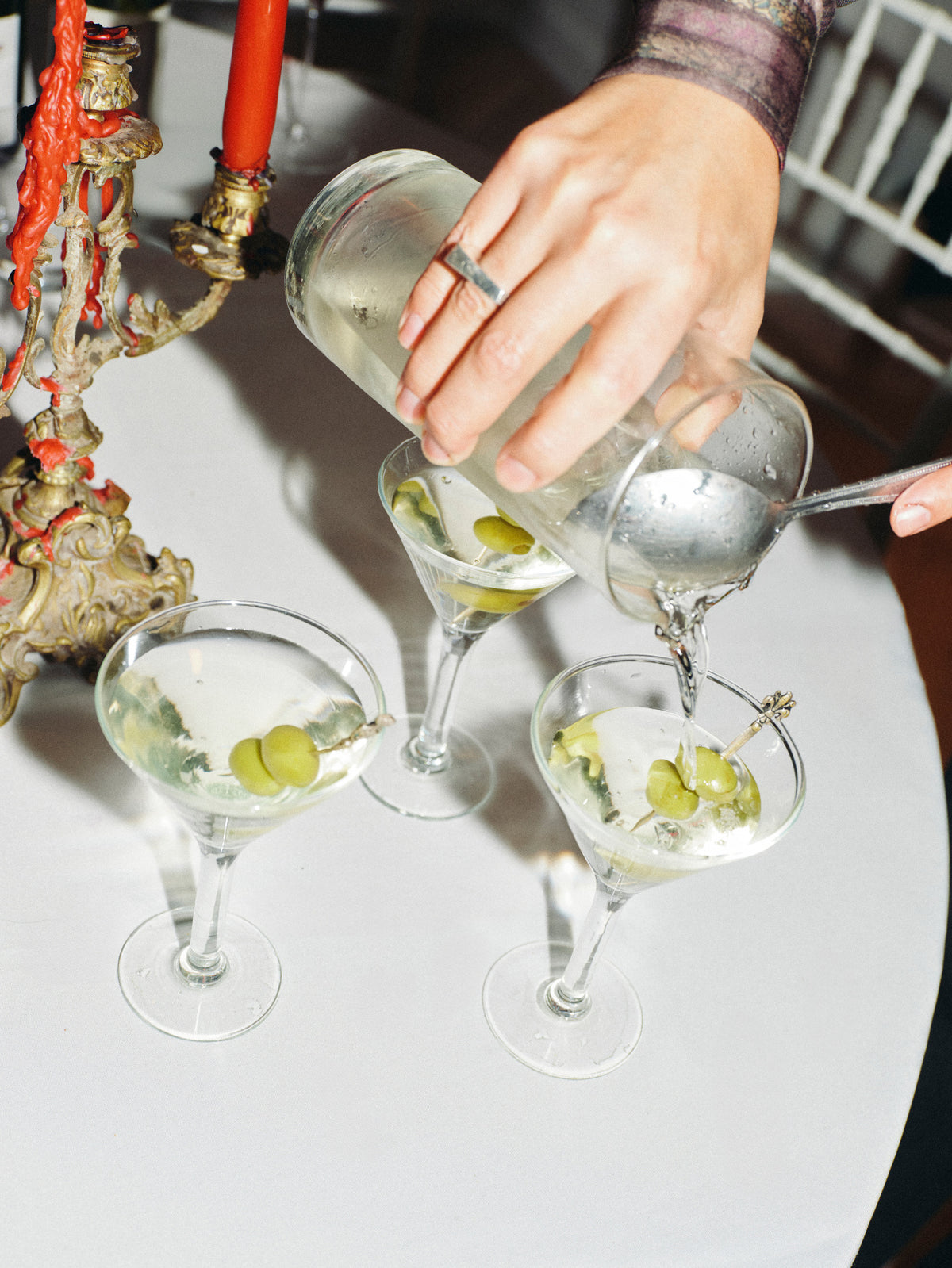 A hand pouring a clear liquid from a glass bottle into three martini glasses containing white wine, olives, and garnishes, set on a white table with an ornate candelabra in the background.