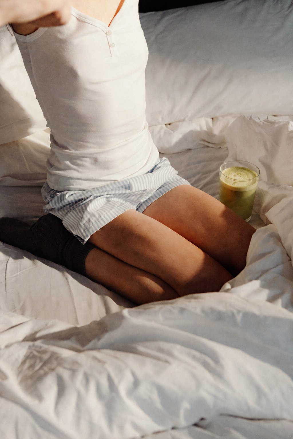 A person kneeling on a bed wearing a white henley top and striped pajama shorts, with a glass of matcha beside them in warm natural light.
